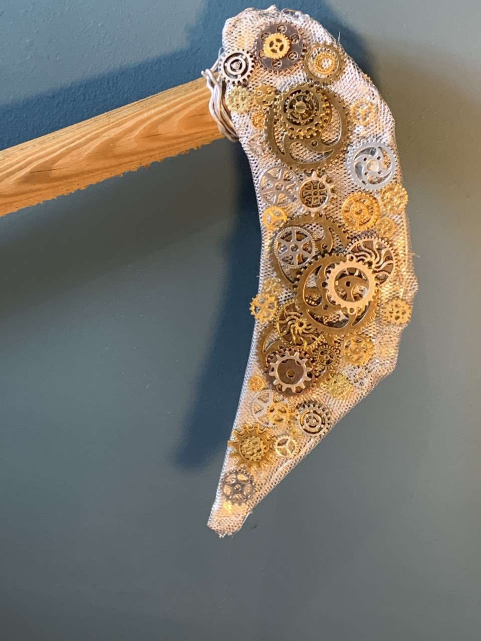 Photo of a 'scythe' with wooden handle and wire 'blade'. The blade is decorated with numerous silver and gold coloured cogs.