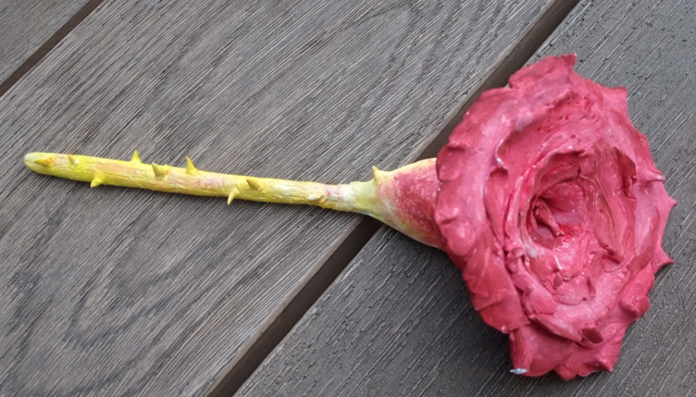 Photo of a clay sculpture of a rose. The stalk is painted gold and looks as though it is rising to cover the red of the rest of the flower.