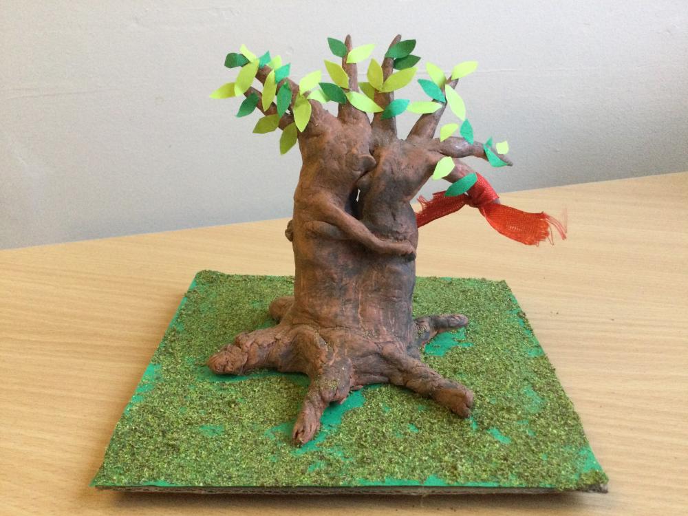Mixed media model of a two-trunked tree, the trunks entwined in an embrace; green paper leaves cling to their branches, and a red scarf or scrap of fabric is tied to one branch.