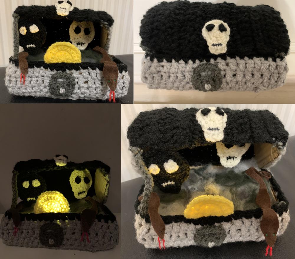 A crocheted Pandora's box, light grey, padlocked, with black lid ajar carrying a skull. Within a black and a white skull and a glowing yellow disc, and a swirl of white and grey wool. Two brown felt snakes with forked red tongues and blue eyes slither out