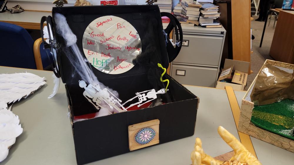A black box, open, 'daisy' or 'sun' symbol on the front; inside the box are skeletons, snakes, spiders' web and a circle on the lid of the box mentioning Greed, Pain, War, Sickness, Guilty, Loss, Jealousy, Evil, Death, Anger—all in red—and in green, Hope