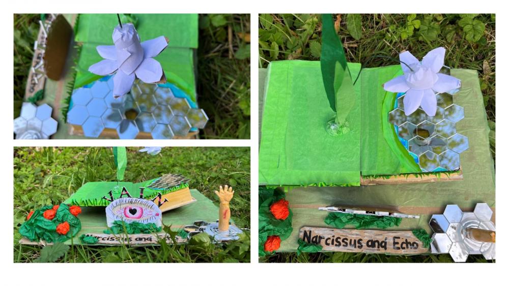 A model made from cardboard and tissue paper, showing a Narcissus flowering growing from an open book, drooping over its own reflection in a pool created from mirrors. Nearby a tissue paper plant flowers, and a small hand reaches up out of another pool.