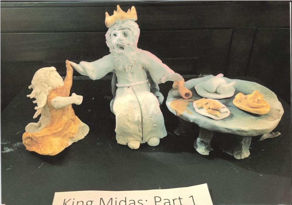 A clay model of Midas at the dining table eating food that is beginning to turn to gold. His daughter has just run to him and is in the process of turning to gold.